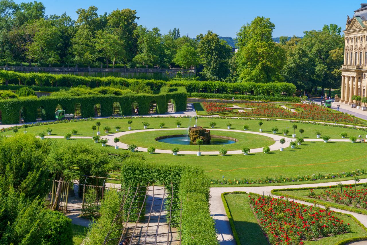 A scenic view of a formal garden featuring a circular fountain, lush greenery, and vibrant flower beds on a sunny summer day.